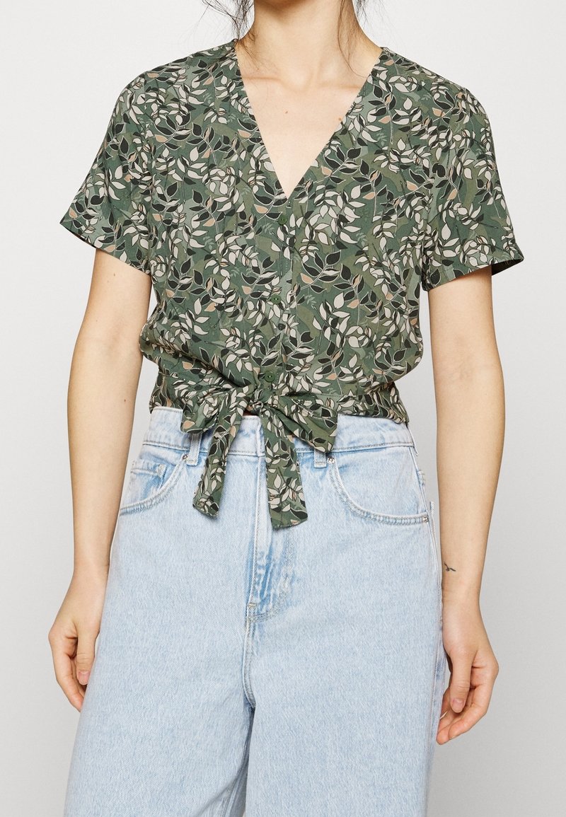 Femme portant un chemisier vert à manches courtes, à motif fleuri avec nœud devant, assorti à un jean bleu clair taille haute, debout devant un fond uni.