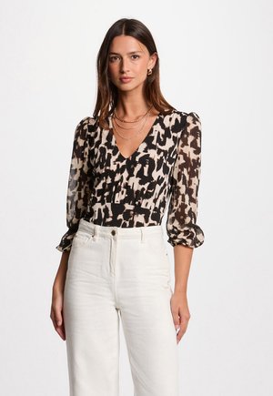 Het model draagt een patroon blouse van doorschijnende stof met pofmouwen, gecombineerd met hoogcilindrische, lichte jeans, terwijl ze poseert met een zelfverzekerde uitstraling.
