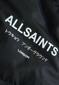 Svart tyg med framträdande vit text: "ALLSAINTS" centrerat, med ytterligare text på japanska och "LONDON" under. Slät textur.