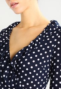 Námořnická halenka s puntíky, která má bílé skvrny, volánkový výstřih a zapínání na knoflíky. Vyrobena z lehkého, hladkého materiálu.