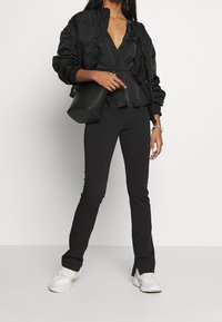 Černá bomber bunda přes černý peplum top, spárována s úzkými černými kalhotami a bílými teniskami. Zahrnuje strukturovanou černou kabelku.