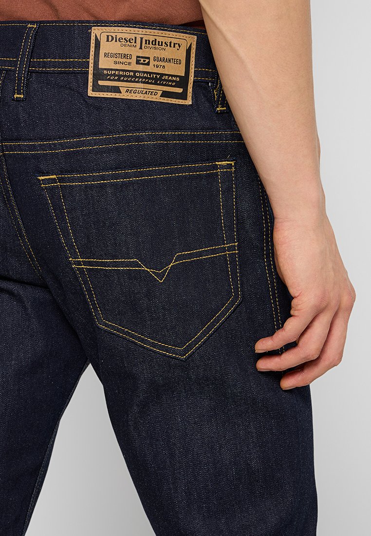 Des jeans en denim foncé présentent un motif de couture jaune, une étiquette en cuir de marque à la taille, et une poche arrière avec un design courbé distinctif.