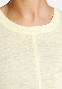 Tecido amarelo-claro, texturizado, com uma qualidade ligeiramente transparente. A parte superior apresenta um decote arredondado e um discreto detalhe de costura vertical.