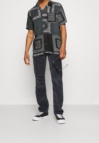 Maglietta nera a maniche corte con motivi di bandana bianca, con bottoni davanti. Pantaloni scuri e sneakers nere con suola bianca. Tiene degli occhiali da sole.