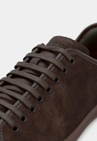 Zapatilla de ante marrón con cordones tonales, ojales redondos y suela de goma lisa. Logotipo grabado en el lateral cerca del talón.