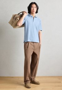 Hellblaues Poloshirt aus strukturiertem Stoff, kombiniert mit braunen gefalteten Hosen und beigen Slippern. Trägt eine tanfarbene Sporttasche.