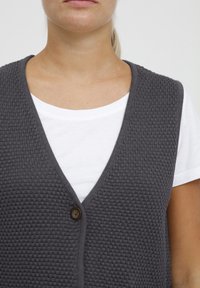 Grijze gebreide vest met een V-hals ontwerp, voorzien van een knoopsluiting aan de voorkant. Draag het over een wit shirt.