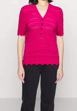 Mujer que lleva una parte superior de manga corta de color rosa brillante con patrones texturizados, botones negros, bordes festoneados y pantalones negros sobre un fondo liso.