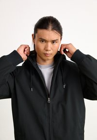 Black jacket with a front zipper and drawstring hood. Features ribbed cuffs and a sleek design, worn by a model with long hair.