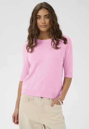 Rozā īss piedurkņu adījuma džemperis ar apaļu kakla izgriezumu, ar ribota auduma apakšu un aprocēm. Valsts kopā ar bēšām augsta jostasvietas biksēm.