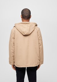 Beige vanntett jakke med hette, som har en jevn tekstur, justerbare snorer og sidelommer. Bakfra viser den en avslappet passform.