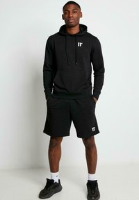 Sudadera negra con cordones y bolsillo, combinada con pantalones cortos negros. Ambos artículos presentan sutiles acentos de logo. Llevado con zapatillas deportivas negras.