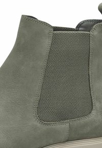 Gros plan sur une botte basse en suède vert avec un panneau latéral élastique texturé et des coutures visibles sur la tige et la semelle.
