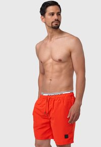 Shorts de bain orange vif avec une taille élastique, un cordon de serrage et un patch noir. Tissu léger avec une texture lisse.