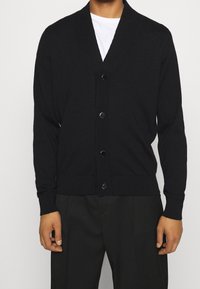 Svart stickad cardigan med V-ringning, framknappar och ribbade ärmslut och nederkant. Slät textur och åtsittande design, bärs över en vit skjorta.