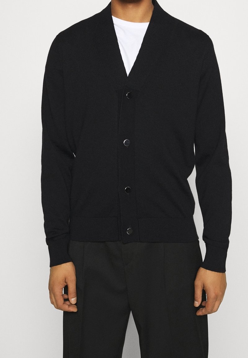 Svart stickad cardigan med V-ringning, framknappar och ribbade ärmslut och nederkant. Slät textur och åtsittande design, bärs över en vit skjorta.