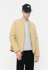 Beige dunjakke med glidelås, grå t-skjorte, svart caps og blåtonet solbriller. Har merking på jakken og capsen.