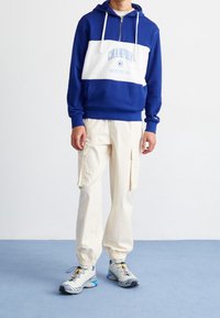 Niebieska i biała bluzka z kapturem zapinana na zamek z napisem "CHAMPION", beżowe spodnie cargo oraz szare buty sportowe z żółtymi i niebieskimi akcentami.