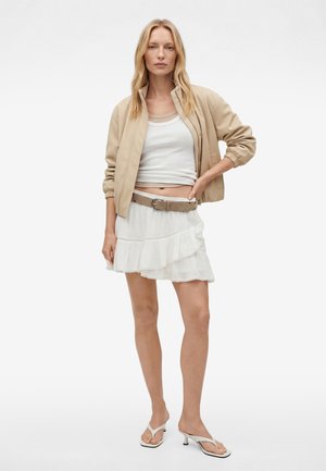 Blonde Frau trägt beige Bomberjacke, weißes Tanktop, geschichteten weißen Rüschrock mit beigen Gürtel und weiße Sandalen mit Absatz, steht vor weißem Hintergrund.