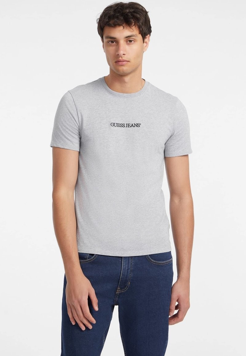 Sivá bavlnená tričko s krátkymi rukávmi, okrúhlym výstrihom a čiernym logom "GUESS JEANS" na prednej strane, nosené s tmavomodrými džínsami.