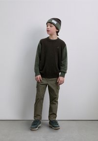 Pull en maille marron et vert à manches longues, pantalon cargo olive et baskets vertes. La tenue est complétée par un bonnet avec un détail d’étiquette.