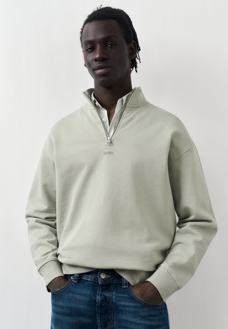 Νέος άνδρας με σκουρόχρωμο δέρμα και κοντά dreadlocks που φοράει ανοιχτό γκρι φούτερ με φερμουάρ έως το στήθος και μπλε τζιν, με τα χέρια στις τσέπες, ουδέτερο φόντο.