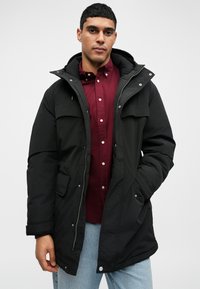 Parka preta com fecho de zíper, bolsos frontais duplos, capuz ajustável e um corte descontraído, usada sobre uma camisa bordeaux.