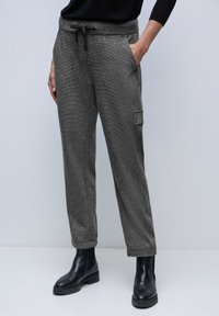 Personne portant un pantalon gris texturé avec des poches avant et latérales, une taille noire à cordon, des bottines noires et un haut à manches longues noir.