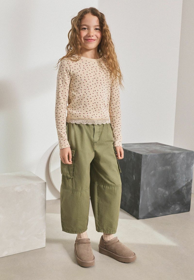 Smilende pige med langt krøllet hår bærer en beige blomsterlangærmet top, grønne brede cargobukser, beige sokker og brune slip-on sko.