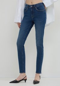 Jean bleu foncé slim fit portés avec des escarpins à talons kitten noirs et une chemise blanche oversize, montrés de la taille aux pieds.