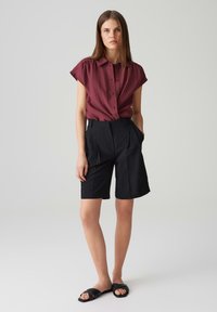 Bordeaux rood kortemouwen overhemd met knopen en geplooide schouders, gecombineerd met op maat gemaakte zwarte korte broek en zwarte slippers. Soepele stof, relaxte fit.