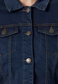 Mörk denimjacka med knäppning framtill, två bröstfickor, kontrasterande gul sömnad och metallknapp-accenter.