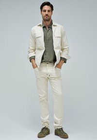 Chaqueta beige clara con dos bolsillos en el pecho sobre una camisa verde apagado, combinada con pantalones crema con cordones y zapatillas verde oliva. Diseño minimalista.