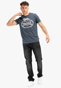 Camiseta de cuello redondo azul marino con un gráfico blanco de un león y texto. Llevada con jeans negros y zapatillas blancas. Ajuste casual, textura suave.