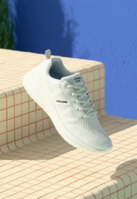 Bijela Jack & Jones tenisica s pletenim uzorkom i vezicama na bež pločicama, plava zidna pozadina i sjena zelenog lišća.