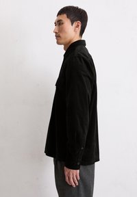 Giovane uomo in camicia di velluto nero e pantaloni grigi in piedi di lato contro una parete bianca semplice.