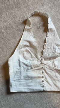 White halter crop top with scalloped edges, embroidered floral eyelet pattern, and covered fabric buttons down the front, laid on gray fabric.