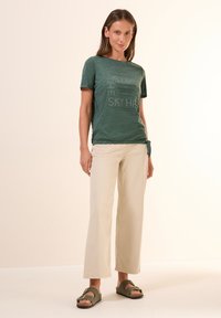 T-shirt vert texturé avec texte imprimé, associé à un pantalon large beige clair et des sandales vertes. Le T-shirt présente un détail de nœud sur le côté.