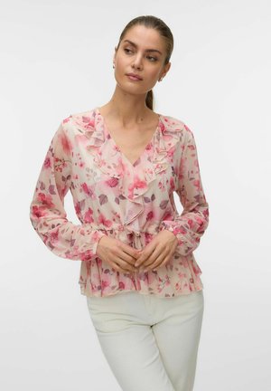 Kobieta w jasno różowej, kwiatowej bluzce z falbankami i długimi rękawami, ubrana w białe spodnie, stojąca na jednolitym tle.