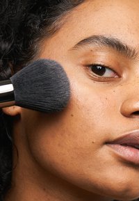 Svart sminkpensel med en tät, mjuk borsthuvud som applicerar produkt på en kind. Hudens textur visar fräknar och en naturlig läppfärg.