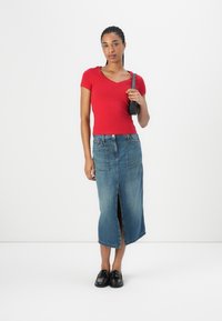 Haut rouge à manches courtes et décolleté en V associé à une jupe midi en denim avec une fente devant. Le modèle porte des mocassins noirs et tient un petit sac noir.