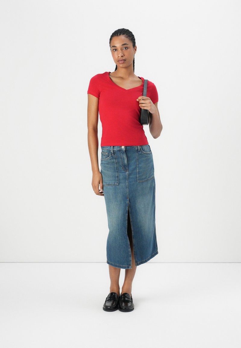 Haut rouge à manches courtes et décolleté en V associé à une jupe midi en denim avec une fente devant. Le modèle porte des mocassins noirs et tient un petit sac noir.