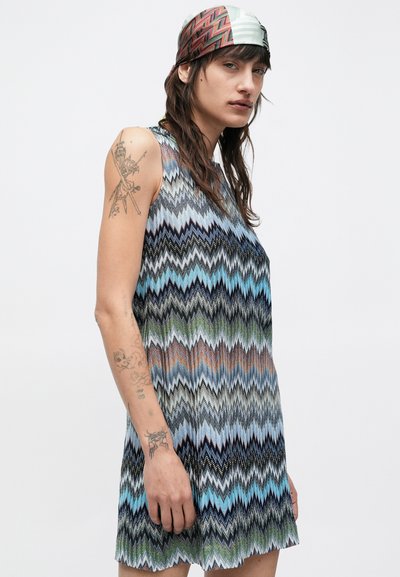 Osoba s dlhými tmavými vlasmi oblečená v šatách bez rukávov s potlačou a farebnom šatke na hlave, ktorá ukazuje tetovania na ramene, stojaca na bielom pozadí.