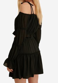 Abito nero off-the-shoulder con maniche lunghe, texture arricciata, polsini svasati e gonna a strati. Realizzato in un tessuto leggero e fluido.