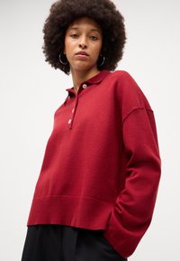 Pólo vermelho de manga longa com colarinho, três botões, bainha canelada e corte descontraído. Feito de um tecido macio e texturizado.