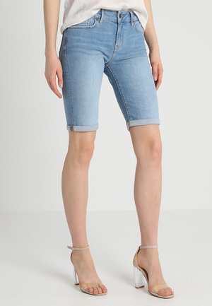 Frau trägt hellblaue Jeans-Shorts bis zum Knie mit umgeschlagenem Saum und beige High-Heel-Sandalen mit transparenten Riemen auf weißem Hintergrund.
