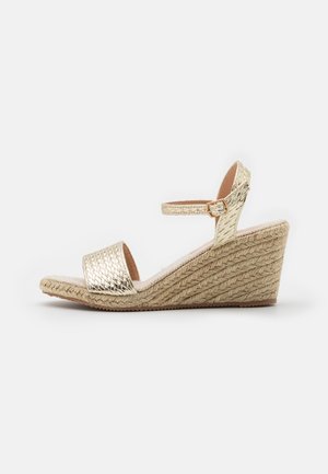 Guldmetallic kile-sandal med vævet juteplatform, ankelrem og åben tå på en ens hvid baggrund.