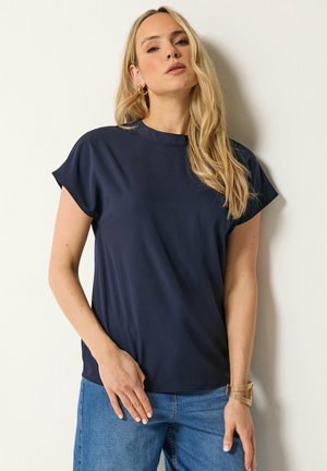 Kobieta z długimi blond włosami, ubrana w granatowy krótki top z krótkim rękawem, niebieskie dżinsy, złote kolczyki-koła oraz złotą bransoletkę, stoi przy białej ścianie.