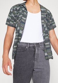 Camisa de manga corta en tejido verde oscuro con patrones geométricos en blanco y azul. Llevada sobre una camiseta sin mangas blanca y combinada con pantalones vaqueros grises.