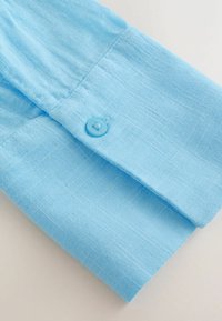 Manche de chemise en coton bleu clair avec une texture subtile, ornée d'un bouton bleu sur le poignet et de bords soigneusement cousus.